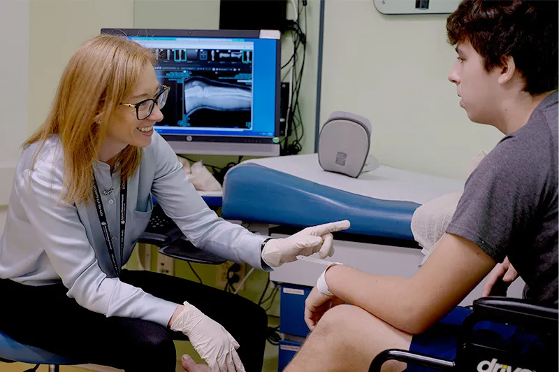 Woman clinician wearing glasses and robber gloves points at a male patient's leg.