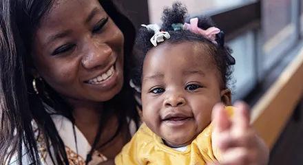 A black mother smiles down at her baby who smiles and points at the camera.