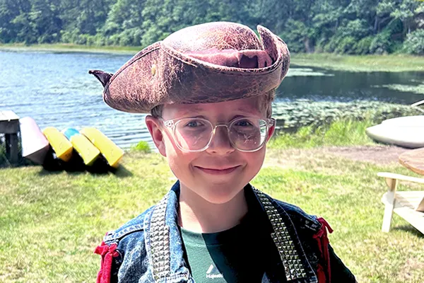 Boy wearing hat and glasses stands at edge of pond