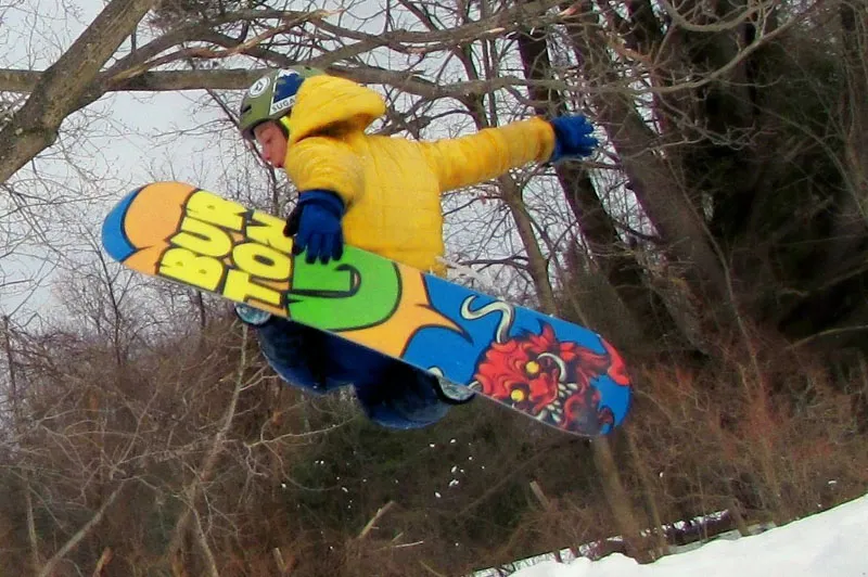 Teen boy flies through the air while riding his snowboard in snowy, wooded area