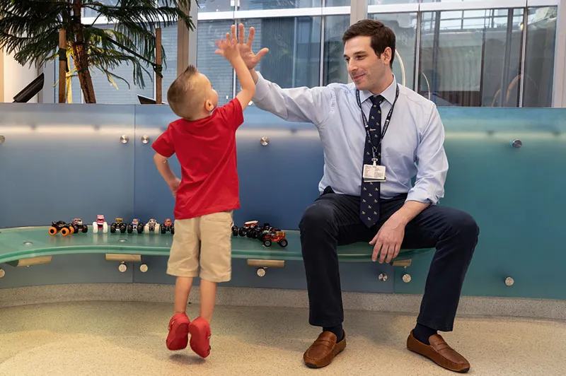 Doctor seated on bench gives high-five to young boy standing on his tippy toes