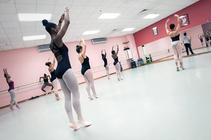 A group of ballet dancers stand in a dance studio with their hands up over their heads.