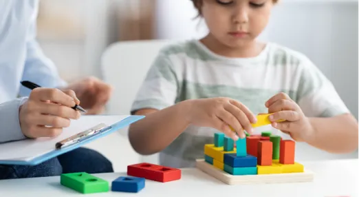 A young child plays with colorful wooden blocks while an adult, holding a clipboard and pen, observes and takes notes.