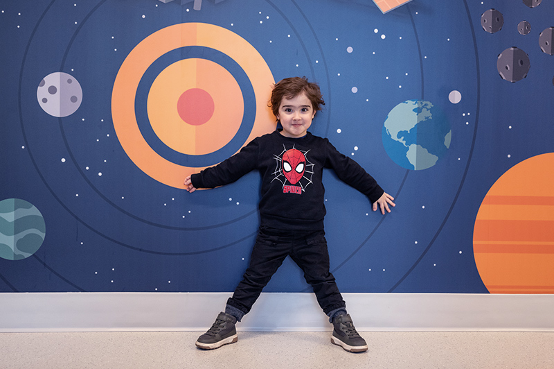 Boy in black outfit stands against a colorfully painted wall with spheres against a blue background