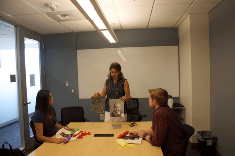 A woman stands in front of a whiteboard holding two images out to a man and a woman who sit at a table.