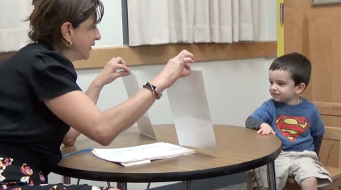 Dr. Choueiri, a woman wearing a black shirt, holds two images up to a toddler boy wearing a superman shirt.